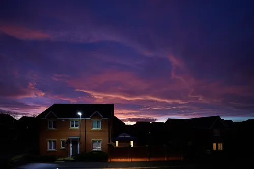 This urban landscape photograph taken in Chesterfield captures a suburban sunset during the evening in the late winter season. The image features a quiet residential street with modern brick houses illuminated by street lights, while nature is present in the background with silhouetted trees against a dramatic sky. The sunset bathes the sky in shades of purple and orange, highlighting the transition from day to night and showcasing the interplay between the urban environment and the natural evening sky.